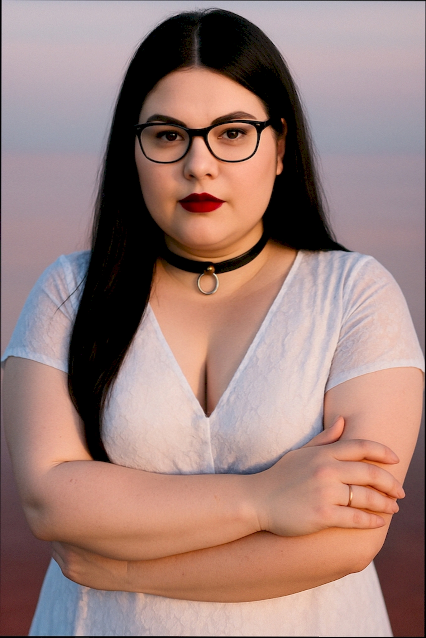 Porträt einer selbstbewussten transfemininen Frau mit schwarzem Haar und Brille, die frontal in die Kamera blickt. Sie trägt ein weißes Kleid mit V-Ausschnitt und ein schwarzes Lederhalsband mit silbernem O-Ring. Ihre Arme sind verschränkt, der Hintergrund ist in sanften Rosa- und Blautönen gehalten. Das Bild vermittelt Entschlossenheit, Selbstbestimmung und stille Kraft. Die Darstellung symbolisiert die Verbindung von Weiblichkeit, Kontrolle und freiwilliger Hingabe.