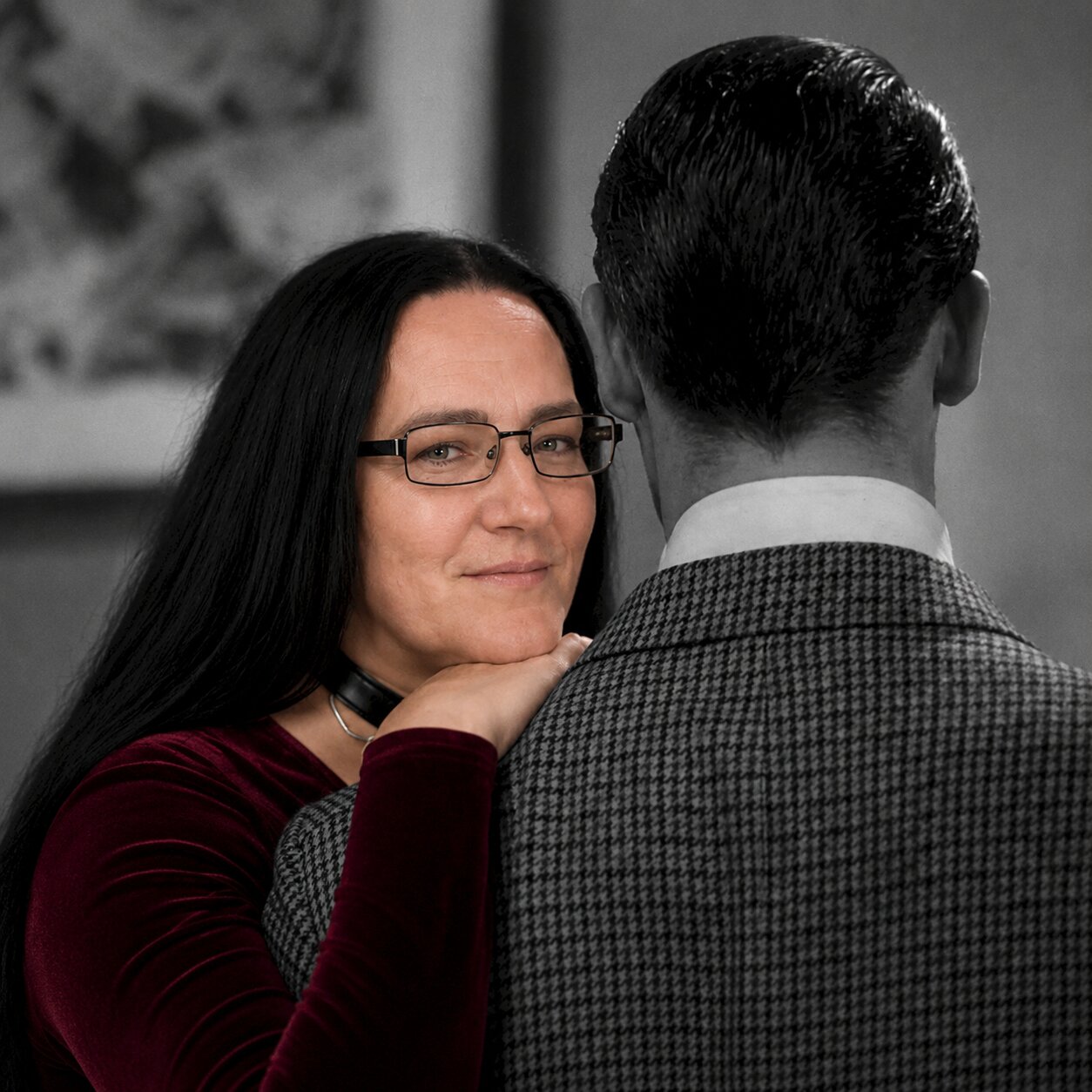 Sarah Jessica Mewes lächelt und legt ihre Hand an den Hals eines Mannes, der ihr den Rücken zuwendet, burgunderfarbenes Samtkleid, schwarze Brille und schwarzes Halsband sichtbar.