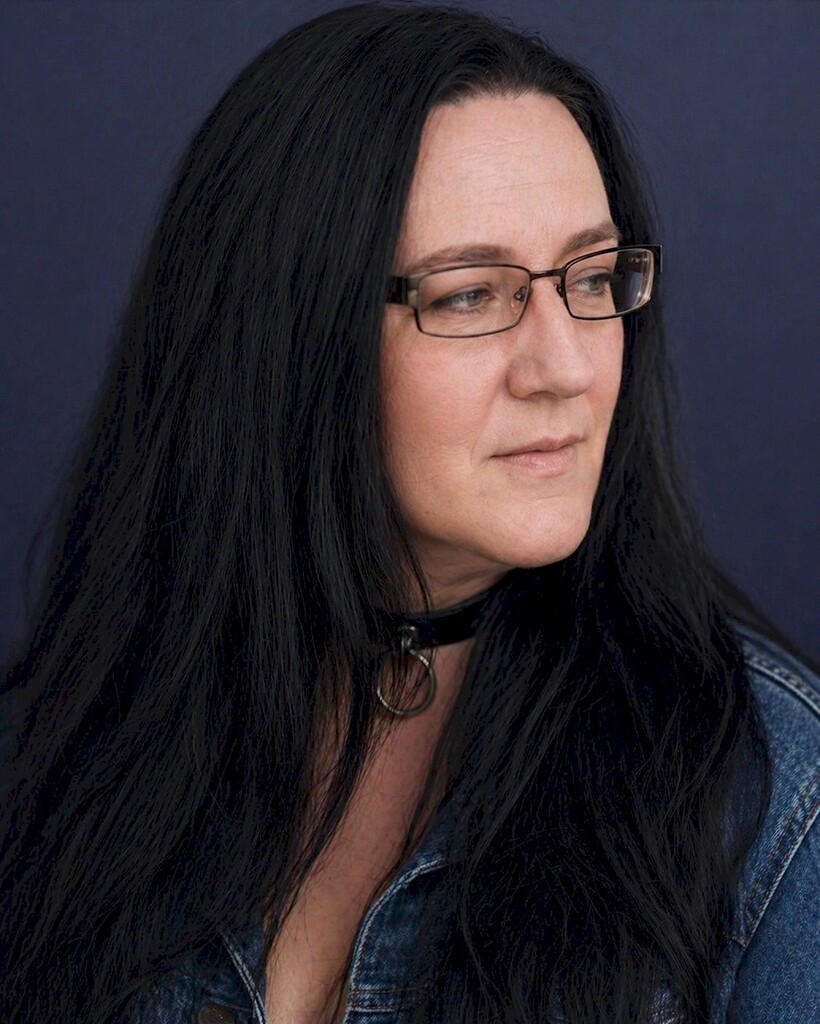 Porträt von Sarah Jessica Mewes mit schwarzem Haar, Brille und schwarzem Halsband vor blauem Hintergrund.